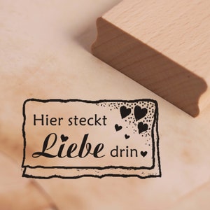 Könnte beinhalten: Ein Holzstempel mit dem Text "Hier steckt Liebe drin" in schwarzer Schrift und einem dekorativen Rahmen. Der Stempel enthält Herzillustrationen. Der Griff des Stempels ist oben rechts sichtbar.