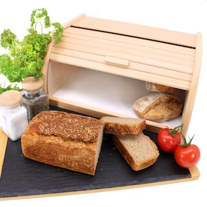 Puede incluir: Una caja de pan de madera con una tapa con bisagras y un frente de listones. La caja está abierta y contiene varios panes. También hay algunos tomates y hierbas en una tabla de cortar de pizarra delante de la caja.