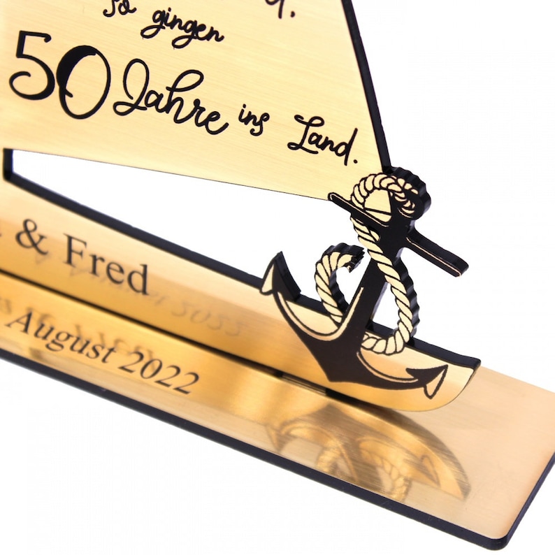 May include: A gold-colored wooden sailboat with a black anchor and rope. The sailboat has the text "50 Jahre ins Land." and "& Fred August 2022" on it.