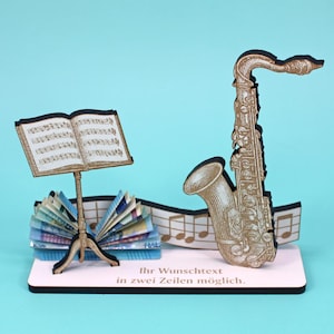 Könnte beinhalten: Dekorative Musikdekoration mit einem goldenen Saxophon, Noten und einem Notenständer. Das Saxophon hat detaillierte Gravuren. Die Noten und der Ständer sind ebenfalls goldfarben. Die Basis ist rosa mit deutschem Text.