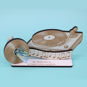May include: A wooden turntable-shaped money holder with a record and musical notes design. The holder has a slot for inserting money, and the text "Ihr Wunschtext in zwei Zeilen möglich" is printed on the base.