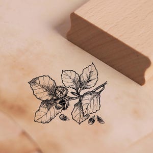 May include: Black and white line art illustration of a branch with leaves and nuts. The image is printed on a wooden block.
