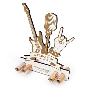 Könnte beinhalten: Ein hölzernes Hochzeitsgeschenk mit einer Gitarre, einem Mikrofon und einer Hand, die eine Rock'n'Roll-Geste macht. Die Worte "JUST MARRIED" stehen auf einem Banner. Die Namen "Oswin & Annegret" und das Datum "29. August 2025" sind ebenfalls enthalten.