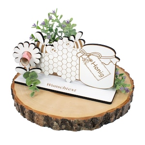 Könnte beinhalten: Weißer Holzhonigtopf mit einem Bienenstock und zwei Bienen, eine weiße Blume mit einem eingerollten Geldschein darin und eine grüne Pflanze auf einer Holzscheibe. Der Topf hat ein Schild mit der Aufschrift "Honig" und die Basis der Holzscheibe sagt "Wunschtext".