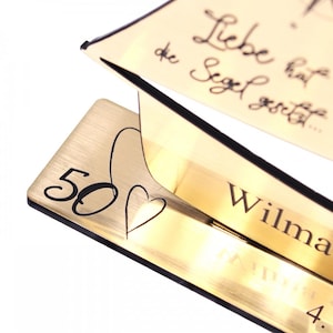 May include: A gold-colored wooden sailboat with the text "Liebe hat die Segel gesetzt..." and "Wilma" engraved on it. The base of the sailboat has the number "50" and a heart engraved on it.
