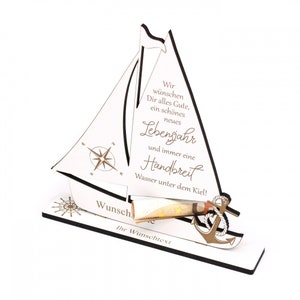 May include: White wooden sailboat with a compass and anchor. The sailboat has a slot for money and a message that reads "Wir wünschen Dir alles Gute, ein schönes neues Lebensjahr und immer eine Handbreit Wasser unter dem Kiel!"  "Wunsch" and "Ihr Wunschtext" are also written on the sailboat.