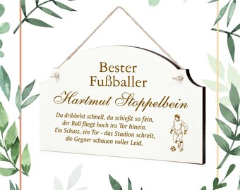 Op de afbeelding: Wit houten bord met de tekst "Bester Fußballer Hartmut Stoppelbein" en een gedicht in het Duits. Het bord heeft een illustratie van een voetballer en is opgehangen aan een natuurlijke touw. Decoratieve groene bladeren omringen het bord.