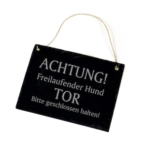 Op de afbeelding: Een zwart leisteen bord met een touw om op te hangen. Het bord heeft de tekst "ACHTUNG! Freilaufender Hund TOR Bitte geschlossen halten!" wat vertaald wordt naar "LET OP! Loslopende hond POORT Houdt gesloten!"