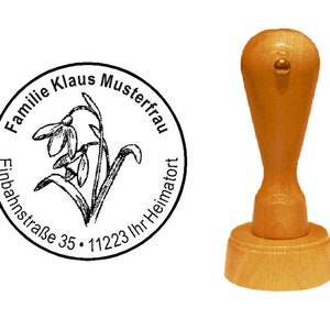 May include: A wooden self-inking stamp with a round design featuring a snowdrop flower, the text "Familie Klaus Musterfrau Einbahnstraße 35 - 11223 Ihr Heimatort", and a wooden handle.