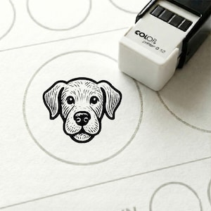 Op de afbeelding: Een zwart-witte hondenstempel op een wit oppervlak. De stempel heeft een cirkelvorm en staat naast een wit en zwart stempelapparaat. Op het stempelapparaat staat de tekst "COLOR printer Q 12".