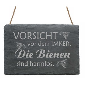 Może przedstawiać: Czarny szyld ze łupka z sznurkowym uchwytem, z napisem "Vorsicht vor dem Imker. Die Bienen sind harmlos." i ilustracją pszczoły.