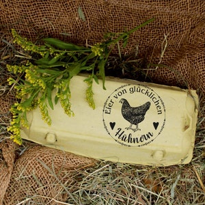 K&ouml;nnte beinhalten: Ein hellgelber Eierkarton mit einer schwarz-wei&szlig;en Illustration eines Huhns und dem Text "Eier von gl&uuml;cklichen H&uuml;hnern". Der Karton ist von Heu und Jute umgeben, mit einem Zweig gelber Blumen.