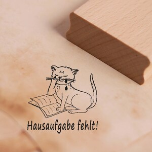 Op de afbeelding: Een houten stempel met een cartoon kat illustratie. De kat zit en huilt terwijl hij een boek leest. De tekst "Hausaufgabe fehlt!" staat eronder.