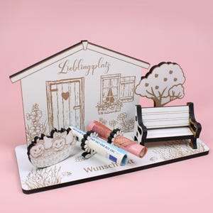 Op de afbeelding: Een decoratieve houten scène met een huis, een bank en een boom. Het huis heeft de tekst "Lieblingsplatz". Opgerolde bankbiljetten worden vastgehouden door decoratieve houders. De basis heeft de tekst "Wunschzeit".