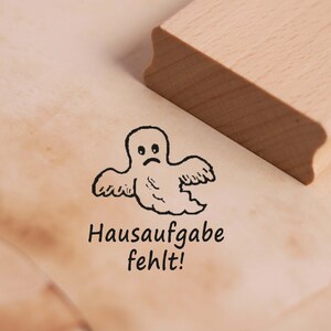 Op de afbeelding: Een rubberen stempel met een cartoon-spook en de tekst "Hausaufgabe fehlt!" wat "Huiswerk ontbreekt!" betekent in het Nederlands.