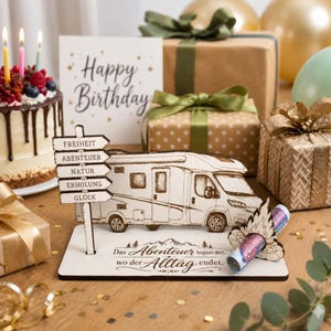 Könnte beinhalten: Ein hölzernes Wohnmobil-Display mit einem Wegweiser, der die Wörter FREIHEIT, ABENTEUER, NATUR, ERHOLUNG, GLÜCK trägt, und einem Sockel mit dem Text Das Abenteuer beginnt dort, wo der Alltag endet, inklusive einer in eine Holzflamme gesteckten Banknote.