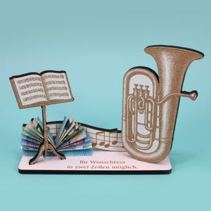 Op de afbeelding: Muziekdecoratie met een gouden tuba en een muziekstandaard. De standaard bevat openstaande bladmuziek. De basis heeft Duitse tekst. Gevouwen papieren elementen completeren het geheel.