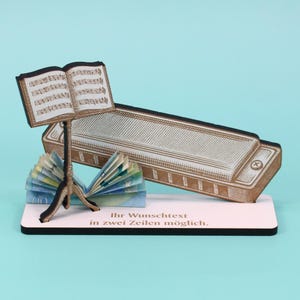 Könnte beinhalten: Eine dekorative Musik-Themen-Anzeige mit einer Miniatur-Mundharmonika, Noten und einem Notenständer. Die Basis hat den Text "Ihr Wunschtext in zwei Zeilen möglich."
