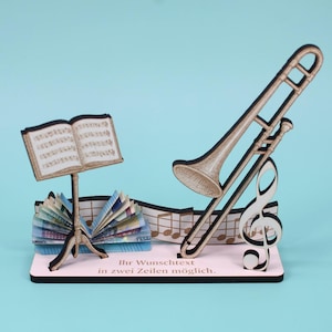 May include: A wooden musical-themed decorative piece featuring a trombone, a treble clef, and a music stand with sheet music. The base is pink with the text "Ihr Wunschtext in zwei Zeilen möglich."