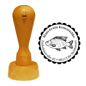 May include: A wooden rubber stamp with a black and white circular design. The design features a fish, the text "Klaus-Erwin Krawutschke", "Einbahnstraße 45", and "00112 Ihr Heimatort".