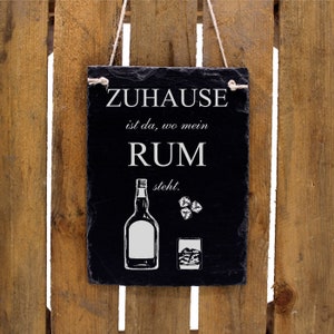 Op de afbeelding: Een zwart leisteen bord met witte tekst die "Zuhause ist da, wo mein RUM steht." luidt. Het bord heeft ook een witte illustratie van een fles rum en een glas met ijsblokjes.