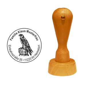 Könnte beinhalten: Ein Holz-Stempel mit einem runden Design, das eine Schwarzweiß-Illustration eines Greifvogels zeigt, der auf einem Fass sitzt. Der Text um den Vogel lautet "Familie Klaus Musterfrau Einbahnstrasse 35 - 11223 Ihr Heimatort".