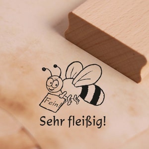 Könnte beinhalten: Eine schwarz-weiße Illustration einer Biene, die einen Zettel mit der Aufschrift "Fein!" hält. Die Biene fliegt und hat einen gestreiften Hinterleib. Der Text "Sehr fleißig!" befindet sich unter der Biene.