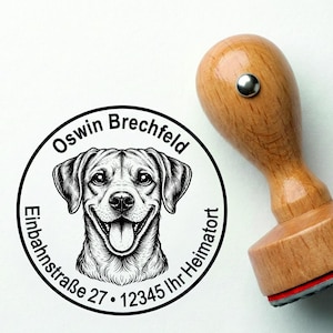 Puede incluir: Un sello de madera con una ilustración de un perro sonriente dentro de un marco circular. El marco incluye el texto "Oswin Brechfeld", "Einbahnstraße 27", "12345 Ihr Heimatort". El sello tiene un mango de madera y una almohadilla de tinta roja.