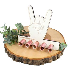 May include: A wooden money holder in the shape of a hand making the rock-and-roll gesture. It features five slots holding rolled-up banknotes. The text "Herzlichen Glückwunsch" is printed on the holder. It rests on a wooden slice, decorated with greenery.