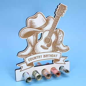 Op de afbeelding: Een verjaardagskaarthouder met countrythema met een cowboyhoed, gitaar en laarzen ontwerp. De woorden "COUNTRY BIRTHDAY" staan op een banner. De houder heeft vijf sleuven voor opgerolde biljetten, tegen een blauwe achtergrond.