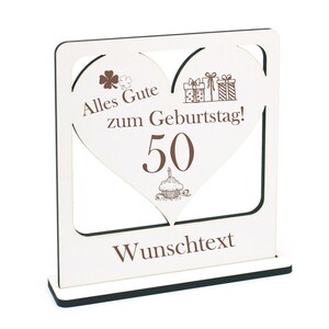 Op de afbeelding: Een wit houten hartvormig bord met een bruine inscriptie die "Alles Gute zum Geburtstag! 50" luidt met een cupcake en een kaars. Het bord heeft een zwarte basis en een zwart frame.