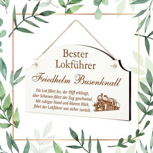 May include: A white wooden sign with brown text that reads "Bester Lokführer Friedhelm Busenknall". The sign has a poem about a train and a small illustration of a train.