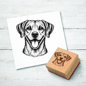 Puede incluir: Ilustración en blanco y negro de la cara sonriente de un perro sobre papel blanco. Un sello de madera con la misma imagen del perro está al lado del papel. El perro tiene la lengua fuera y está sonriendo.