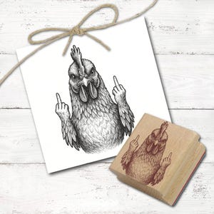 Peut inclure: Illustration en noir et blanc d'une poule faisant un geste obscène, imprimée sur une carte blanche. Un tampon en bois avec la même image est également présent. La carte est attachée avec un nœud en ficelle naturelle.