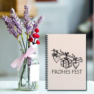 May include: A spiral bound notebook with a beige cover and a black and white illustration of a gift box with hearts hanging from a branch. The text "FROHES FEST" is printed below the illustration.