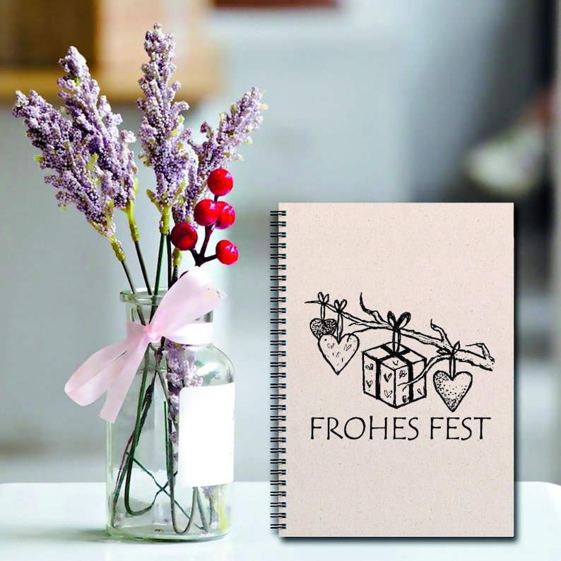 May include: A spiral bound notebook with a beige cover and a black and white illustration of a gift box with hearts hanging from a branch. The text "FROHES FEST" is printed below the illustration.
