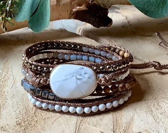Op de afbeelding: Een wikkelarmband met meerdere strengen en een grote, ovale witte steen met grijze aders als middelpunt. De armband is gemaakt met bruin koord en bevat diverse kleine kralen in tinten wit, grijs en metallic brons.
