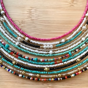 May include: A collection of colourful beaded necklaces, featuring turquoise, white, black, pink, and gold beads. The necklaces are arranged in a fan shape on a wooden surface.