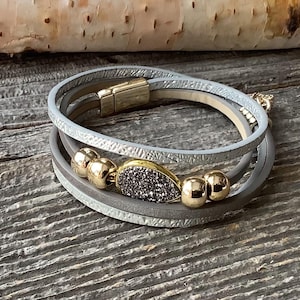 Peut inclure: Un bracelet multi-rangs composé de deux cordons en cuir gris et d'un cordon texturé argenté métallisé. Le bracelet est orné de quatre perles dorées polies et d'une breloque centrale en forme de goutte avec une finition druzy gris foncé.