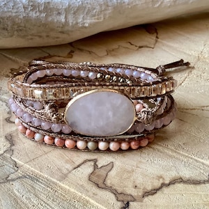 May include: A multi-strand wrap bracelet featuring a large oval rose quartz stone in a gold setting. The bracelet incorporates various pink and gold beads, and is secured with a brown leather cord.