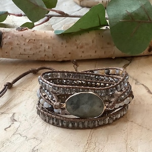 May include: Wrap bracelet featuring a large oval gemstone in a gold setting, surrounded by multiple strands of gray and clear beads. The bracelet is made of brown leather and has a tie closure. The background includes a birch branch and green leaves.