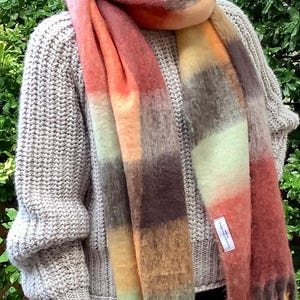 Puede incluir: Un suéter de punto grueso en color gris claro combinado con una bufanda larga de textura suave con un patrón de cuadros grandes en tonos naranja óxido, amarillo apagado, marrón oscuro y verde pálido, con flecos y una etiqueta blanca.