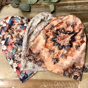 May include: Three soft fabric beanies featuring distinct patterns. One displays a dense floral arrangement in shades of pink, blue, and white. Another shows a light-coloured intricate design. The third features a large, dark-toned mandala print.