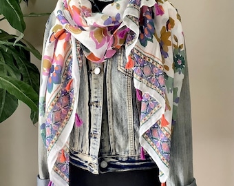 May include: A white scarf featuring a multi-colored floral and geometric pattern with small orange and purple tassels along the edges, draped over a light blue acid-wash denim jacket with silver buttons, displayed on a black mannequin torso.