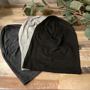 Peut inclure: Un ensemble de trois bonnets souples en noir uni, gris clair et un tissu chiné gris anthracite foncé. Chaque bonnet présente une construction légère et douce avec un sommet arrondi et un bord ourlé simple, conçu pour une coupe décontractée.