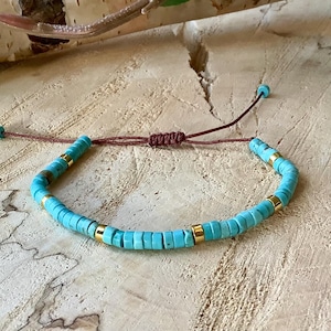 Op de afbeelding: Een turquoise en gouden kralen armband met een bruine koordsluiting. De armband is gemaakt van kleine, ronde kralen met een gouden accent op elke kraal.
