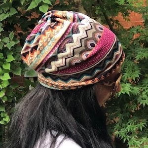 May include: A slouchy beanie hat featuring a multi-coloured geometric pattern with zig-zag lines, stripes, and dotted textures in shades of magenta, orange, teal, brown, and cream. The fabric has a soft, gathered appearance with a wide band.