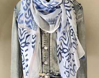 May include: A lightweight white scarf featuring a blue abstract ikat-style pattern, geometric border details, and small blue tassels along the bottom hem, draped over a light-wash denim jacket with metal buttons and a collar.