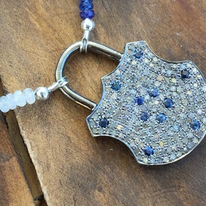 May include: A silver padlock pendant necklace with a shield-shaped design. The pendant is covered in small, sparkling blue and white gemstones.