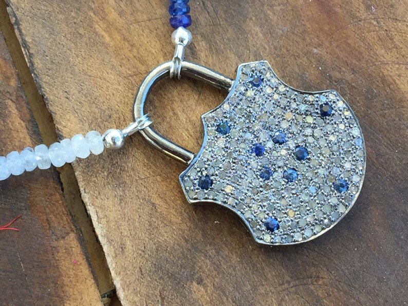 May include: A silver padlock pendant necklace with a shield-shaped design. The pendant is covered in small, sparkling blue and white gemstones.